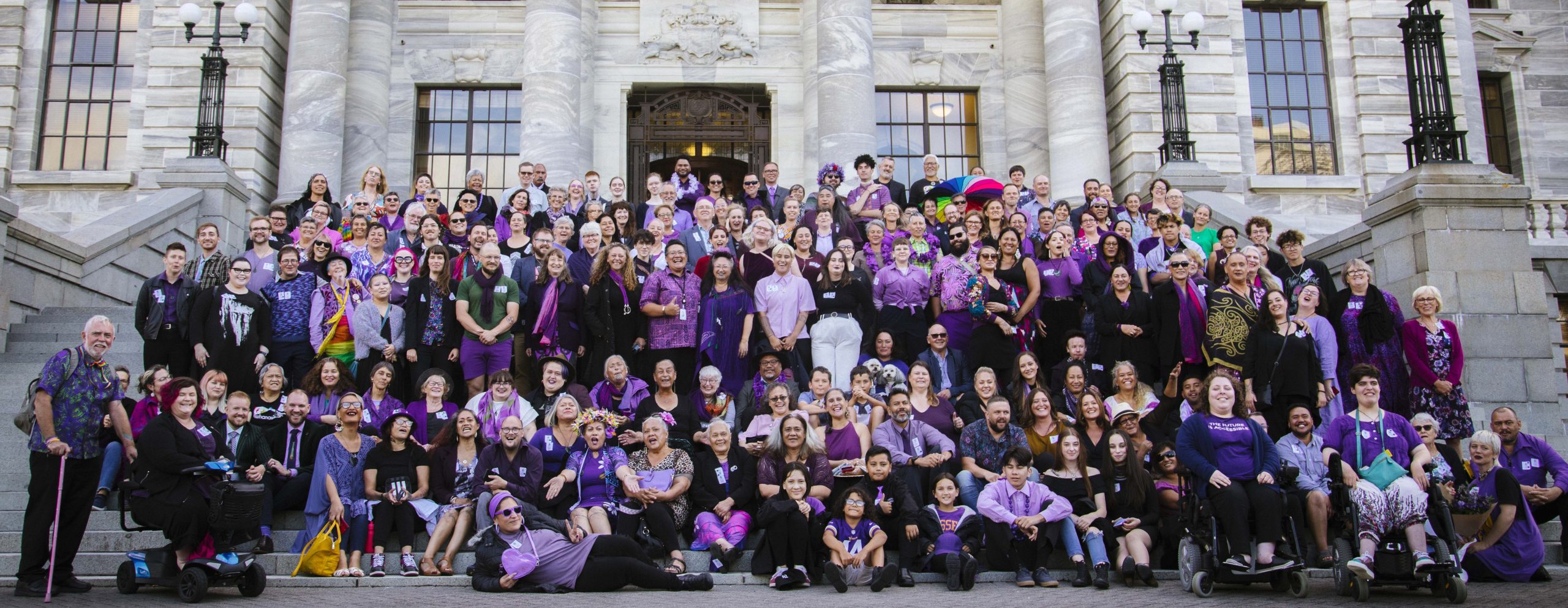 Elizabeth and her whānau, friends, and colleagues on the steps of parliament on the day of her maiden speech. Hundreds of Elizabeth's supporters showed up to "paint parliament purple" - everyone is wearing purple and smiling.