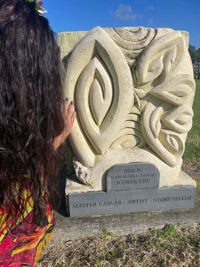 A photo of Elizabeth's father's graveside, featuring a beautiful Māori carving. Elizabeth is photographed from behind, her hand reaching out to touch the carving. 